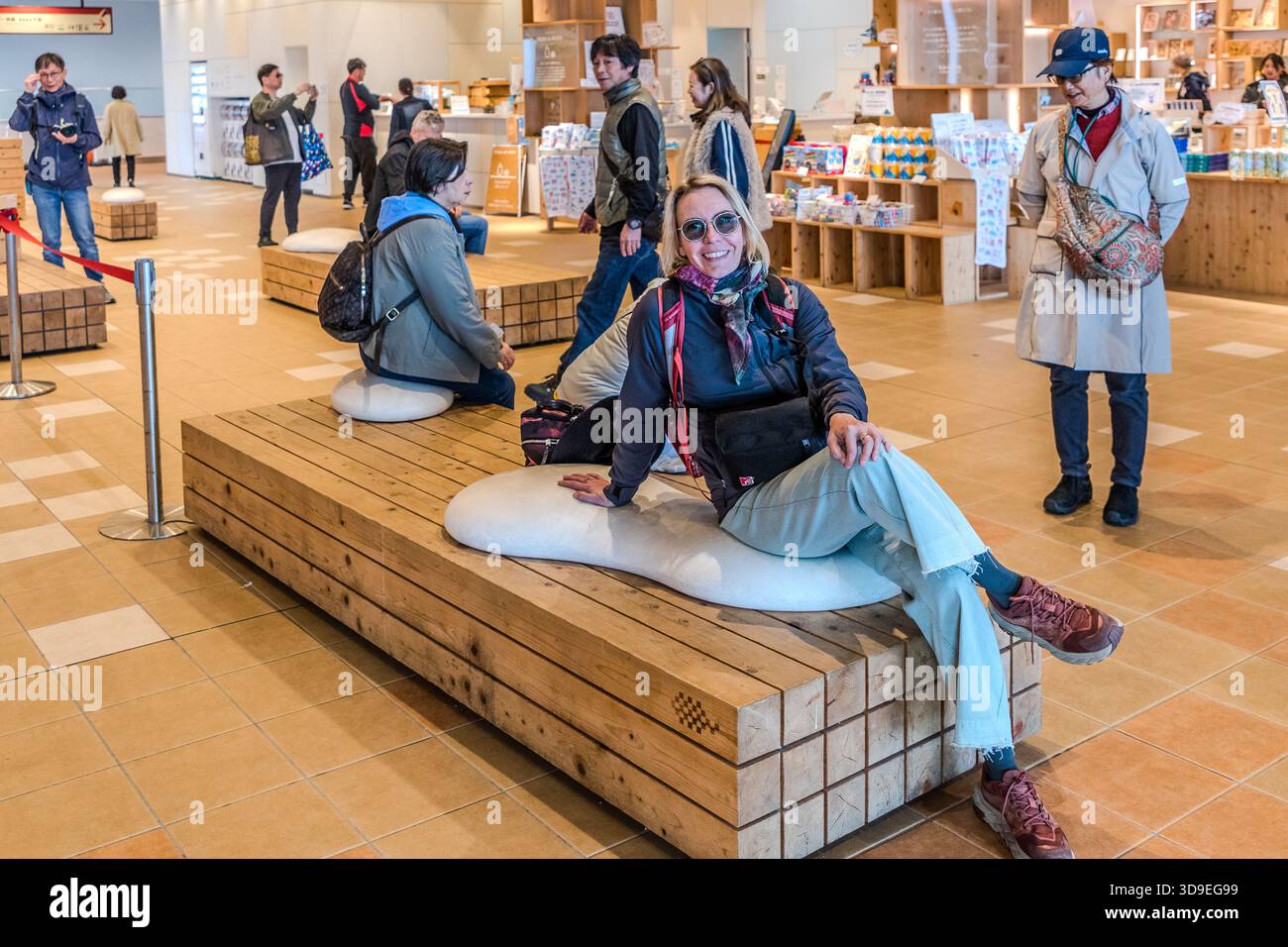 A visitor is sitting on a wooden bench with a white cushion in the entrance hall of a modern visitor centre in Hakone, Kanagawa Prefecture , Japan; in the background is a souvenir shop with other visitors Stock Photo