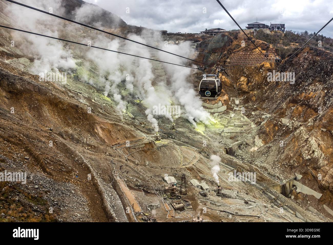A gondola on the Hakone Ropeway crosses the Owakudani Valley (大涌谷, ‘Great Boiling Valley’) in Hakone, Kanagawa, Japan. The area lies within the crater of a volcano that is around 3,000 years old; sulphur deposits (yellow patches) and steam rising from fumaroles are signs of ongoing volcanic activity. The buildings of the Owakudani cable car station can be seen in the background Stock Photo