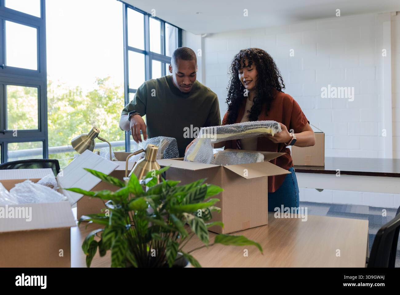 African American coworkers unpacking bubble-wrapped mirrors and brass lamp on table in office Stock Photo