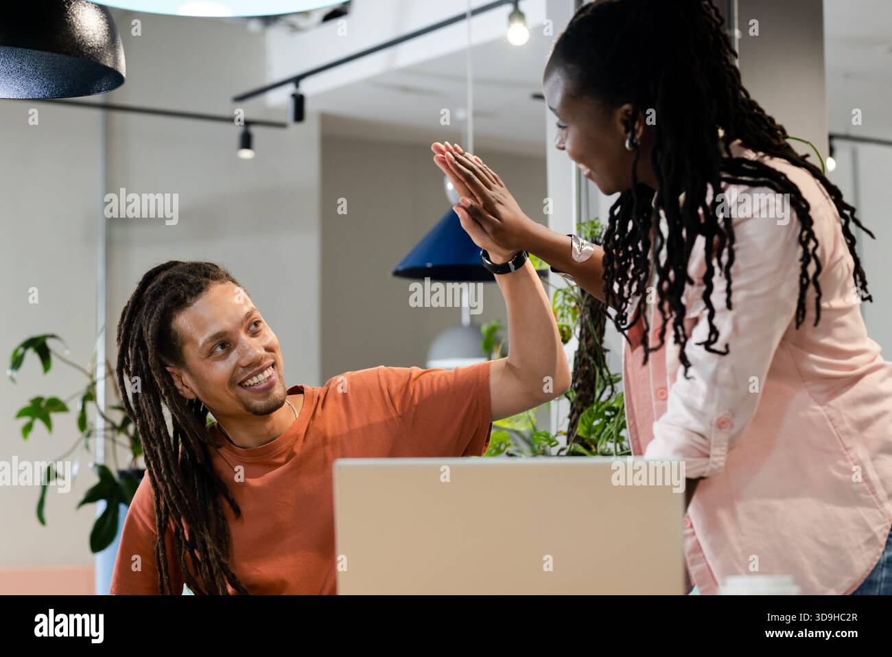 African American coworkers high-fiving over laptop at open-plan office, potted plants Stock Photo
