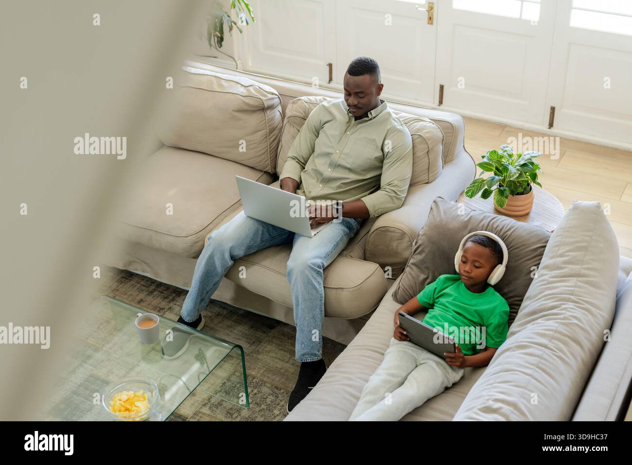 African American father and child sitting on beige sectional at home using laptop and tablet Stock Photo