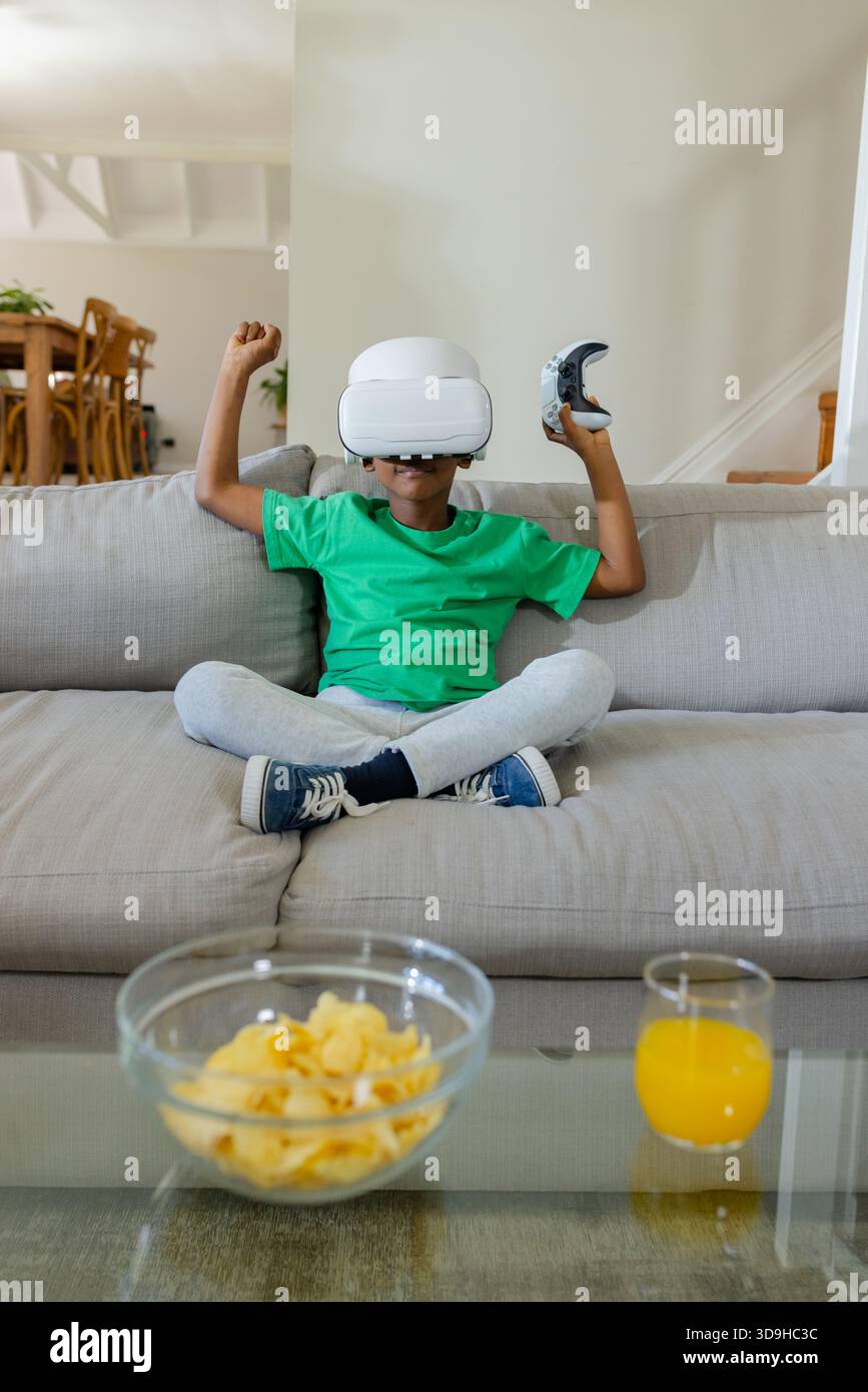 VR headset sitting on light-gray sofa with controller, bowl of potato chips, glass of orange juice Stock Photo