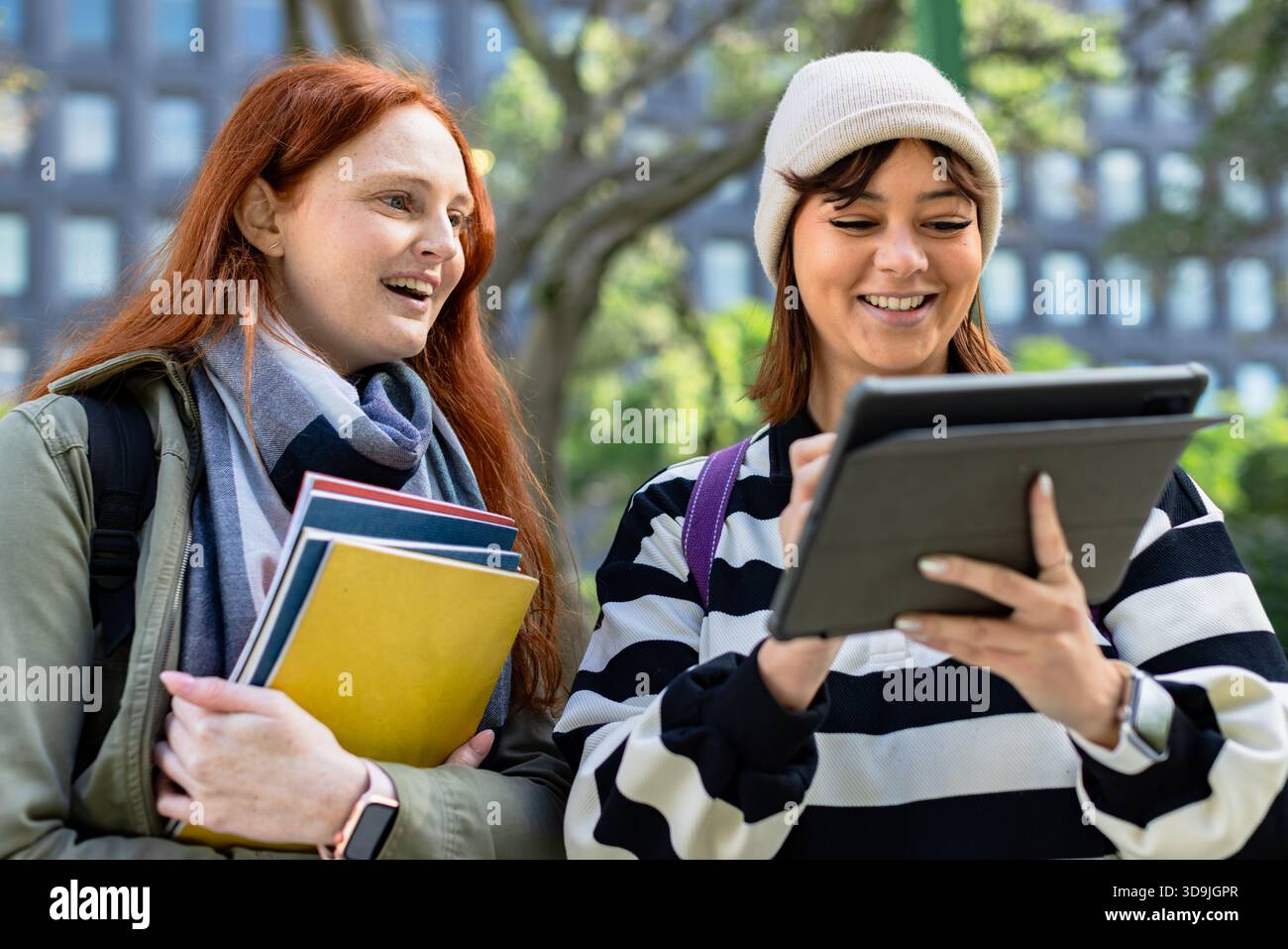 Two female students collaborating on campus walkway, holding notebooks and using stylus tablet Stock Photo