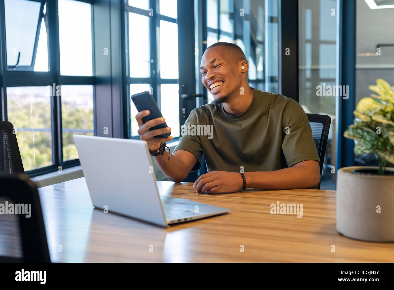 African adult male smiling and checking smartphone screen at conference table with laptop and plant Stock Photo