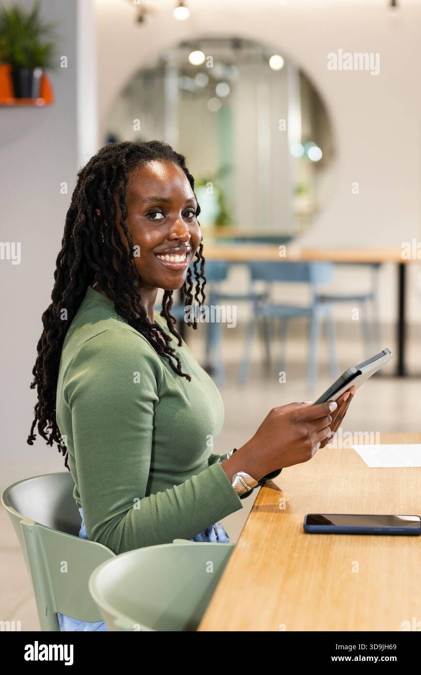 African American woman sitting at coworking table holding tablet smiling with smartphone visible Stock Photo