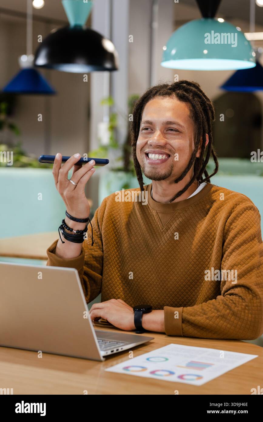 Man smiling and speaking into smartphone while sitting at coworking lounge with open laptop Stock Photo