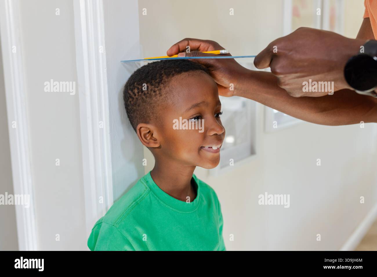 African American father and son measuring height in hallway with book, pencil, boy in green T-shirt Stock Photo