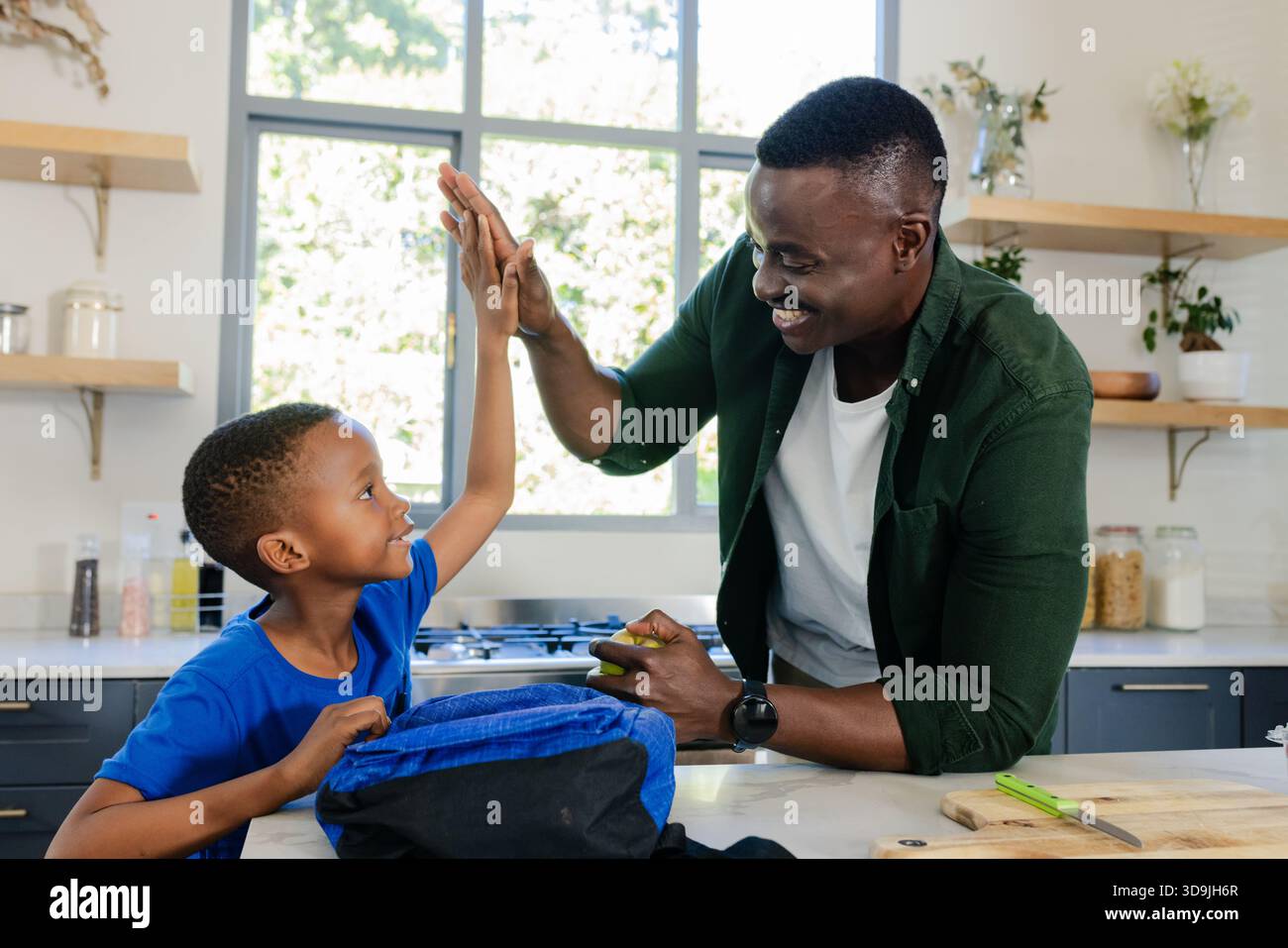 African American father and son high-fiving at kitchen island by window with blue backpack, apple Stock Photo
