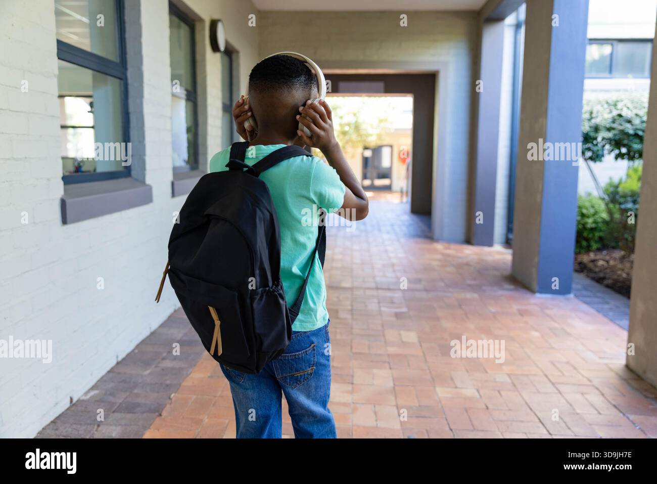 Child is standing in covered corridor on terracotta tiles holding headphones and wearing backpack Stock Photo