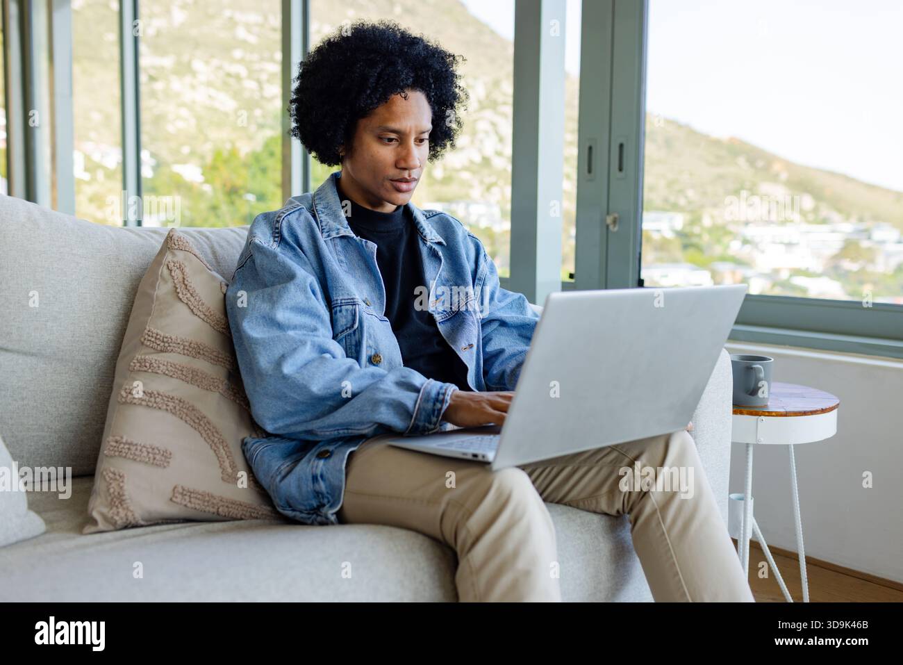 African American man typing on laptop on sofa with coffee mug in living room overlooking hillside Stock Photo