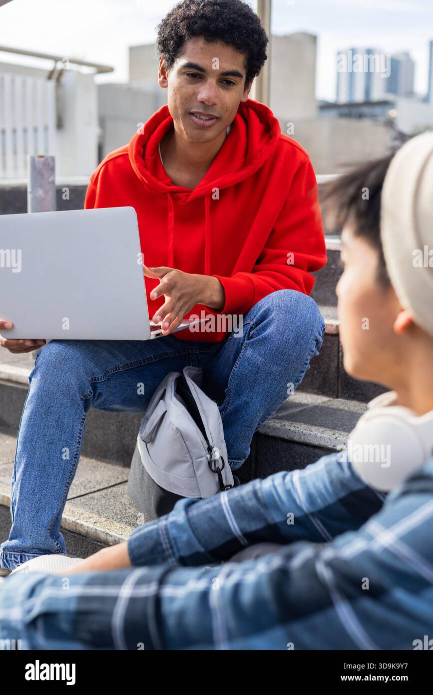 Diverse friends discussing project on laptop while sitting on rooftop steps near gray backpack Stock Photo
