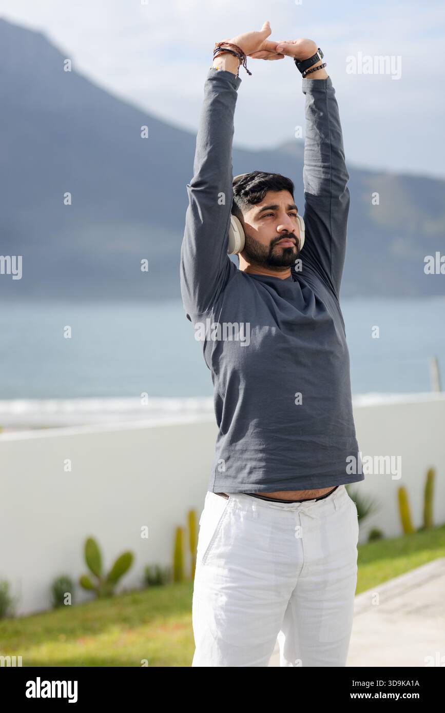 Overhead Man Indian man stretching arms overhead on seaside terrace wearing over-ear ...