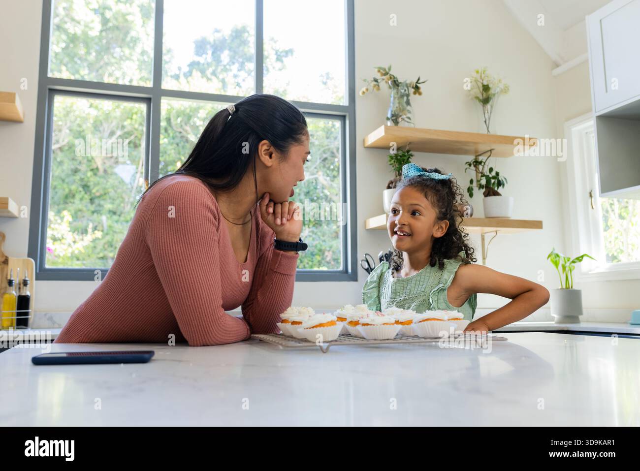 Mother and daughter chatting at marble kitchen island with cupcakes cooling rack and smartphone Stock Photo