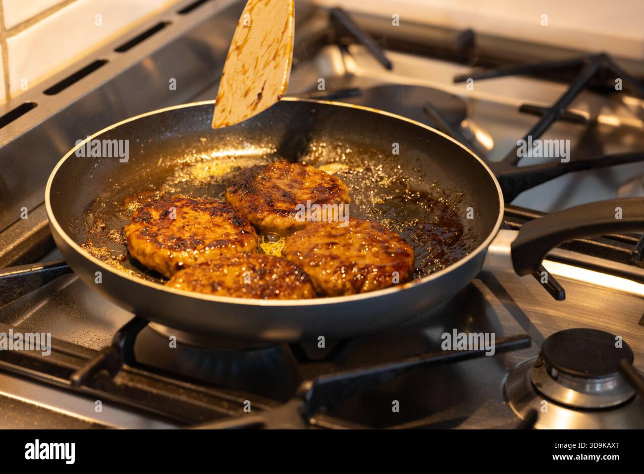 Four seared meat patties are sizzling in non-stick pan on gas stovetop, spatula hovering Stock Photo
