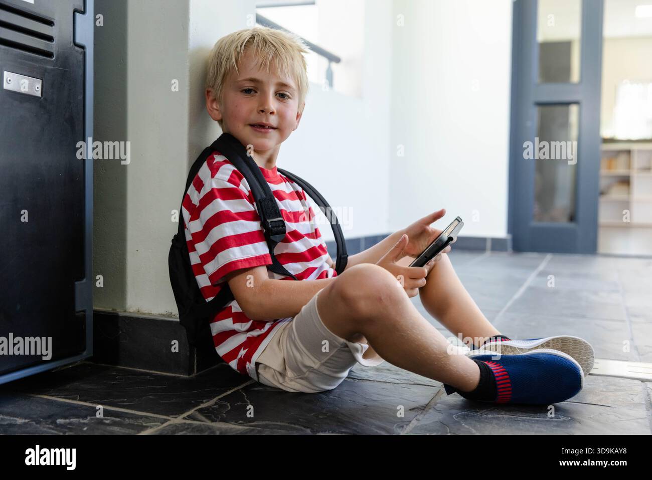 Boy sitting on school hallway leaning on column holding phone and backpack wearing striped tee Stock Photo