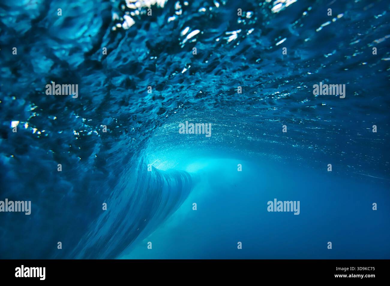 Underwater view breaking ocean wave tunnel deep blue barrel curling above, light filtering through translucent Stock Photo