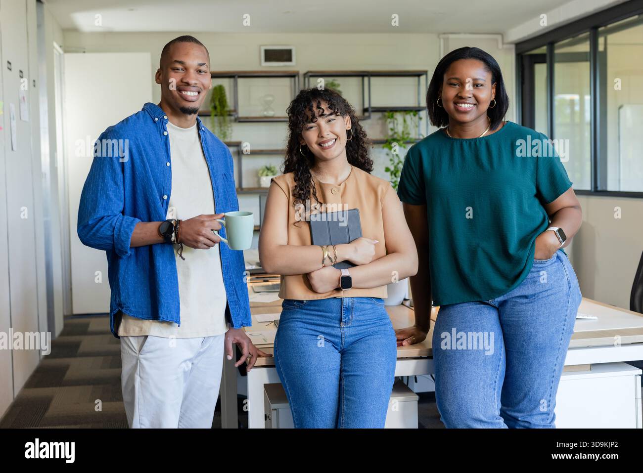 Coworkers posing and smiling in open office at shared desk, holding light green mug and tablet Stock Photo