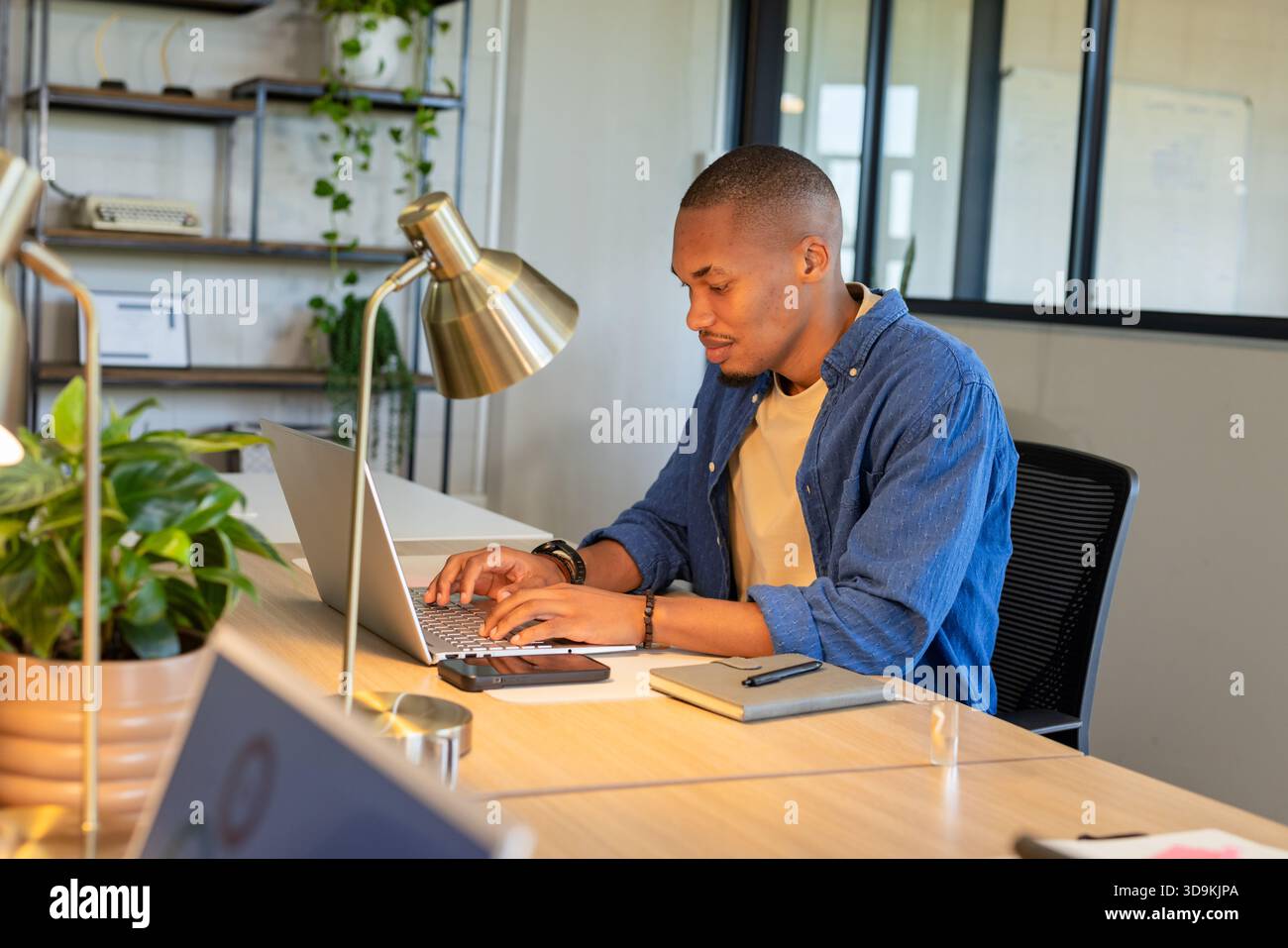 African American man typing on laptop at modern office workspace beside desk lamp and smartphone Stock Photo