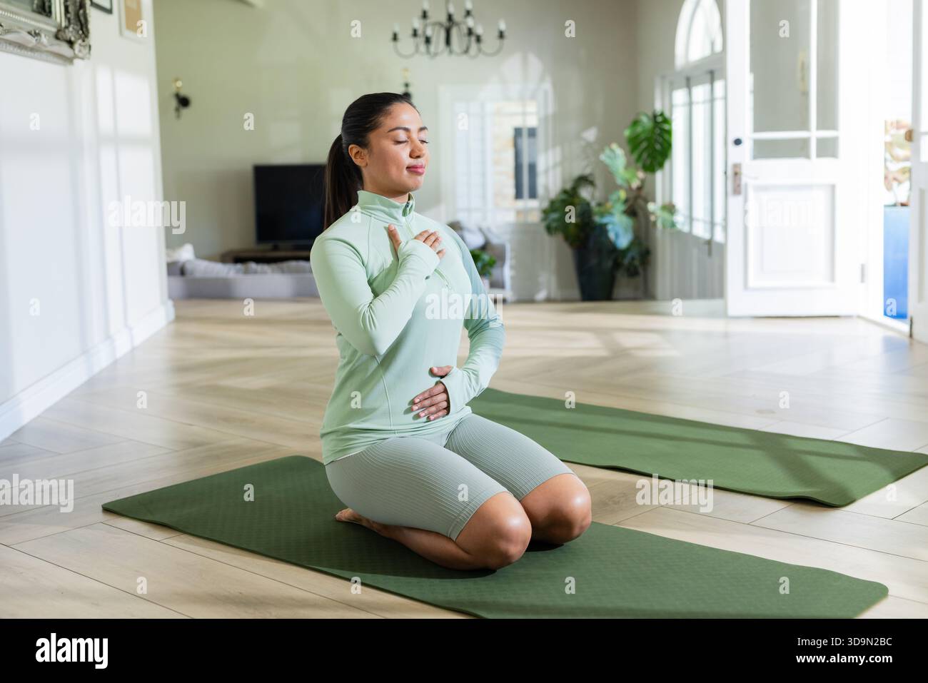Woman kneeling on green yoga mat in bright living room practicing breathwork under black chandelier Stock Photo
