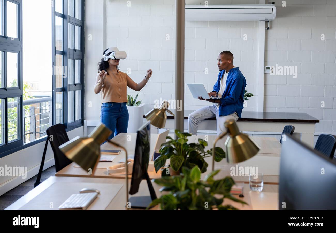 African American coworkers showing VR headset demo while using laptop in casual office with windows Stock Photo