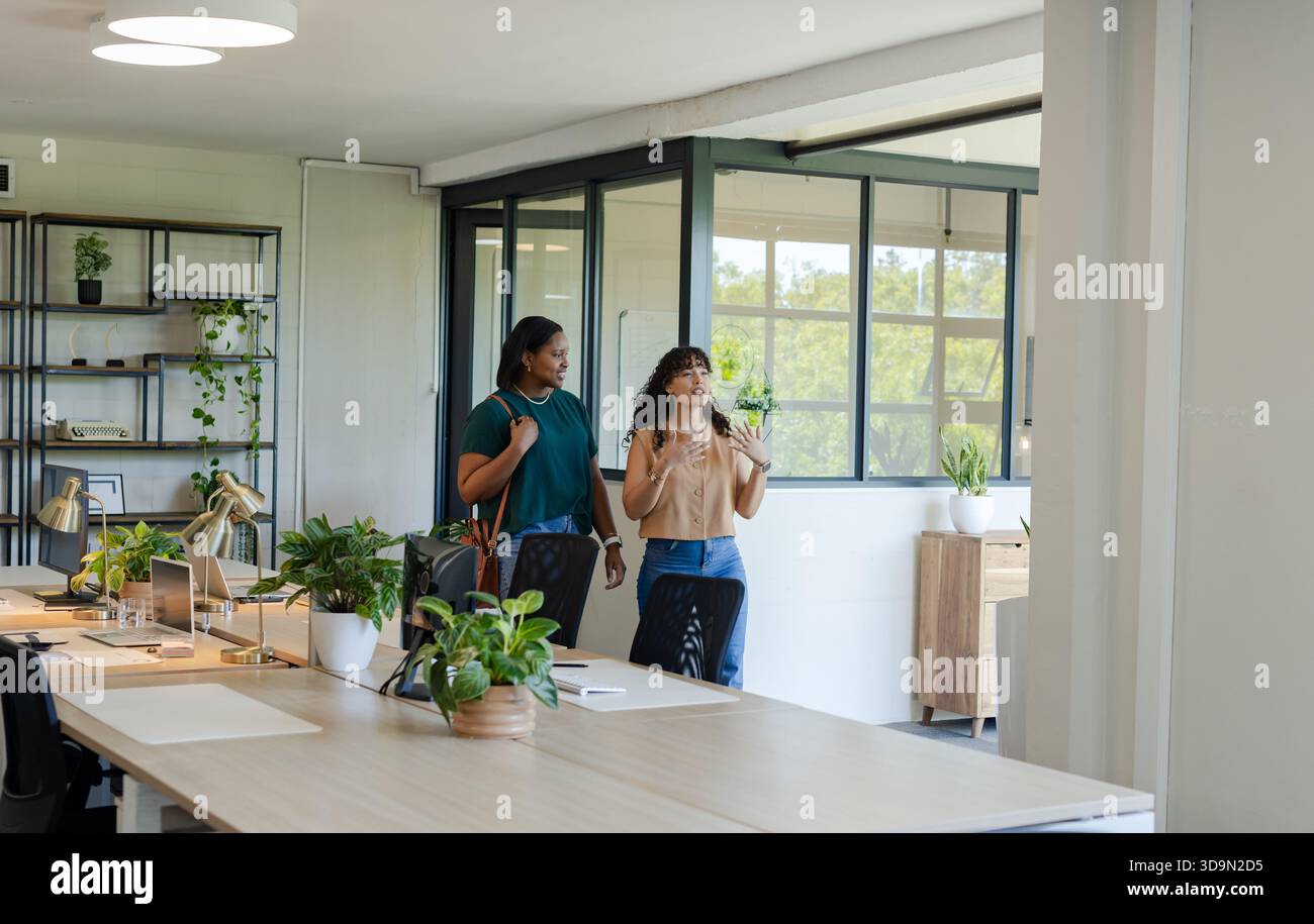Diverse female colleagues walking and discussing in open plan office with wooden desks and laptops Stock Photo