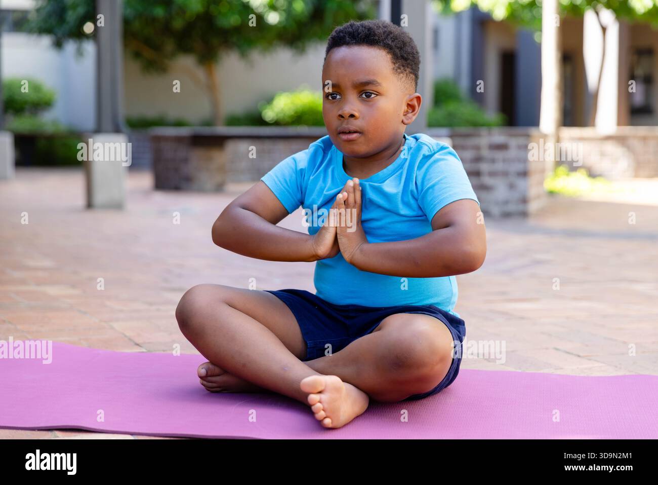 African boy sitting cross-legged on pink yoga mat in courtyard in light blue shirt pressing palms Stock Photo