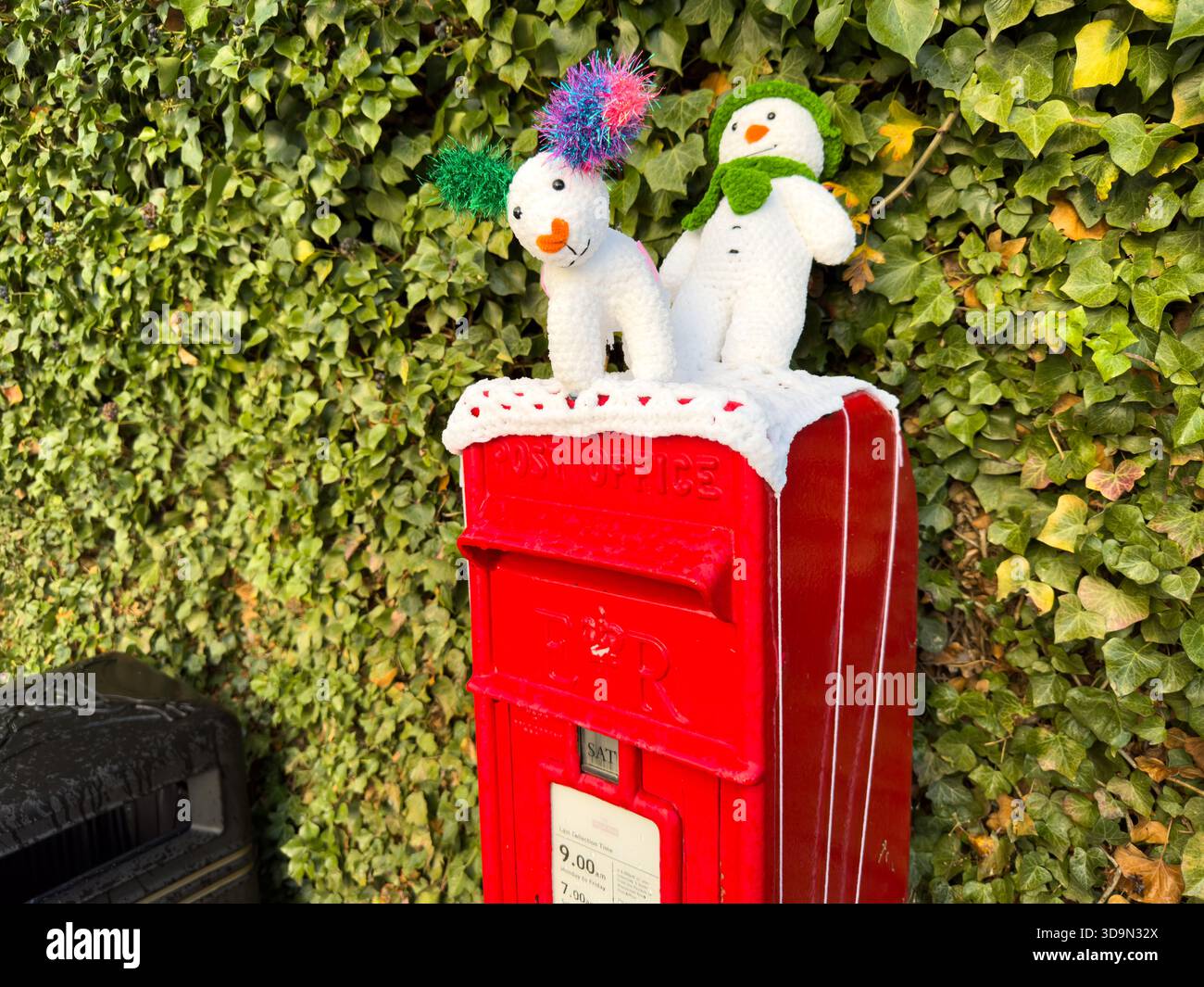 Knitted Christmas figures of a snowman and a reindeer on top of a small red post box on in front of a green leafed hedge on a uk street. - Smartphone Captured Stock Image