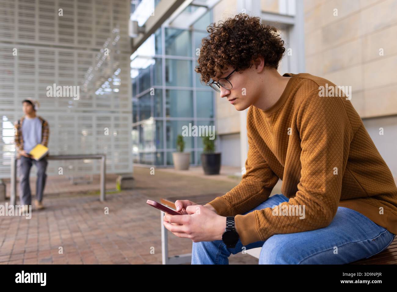 Male students sitting on campus bench using red smartphone with yellow notebook near potted plants Stock Photo