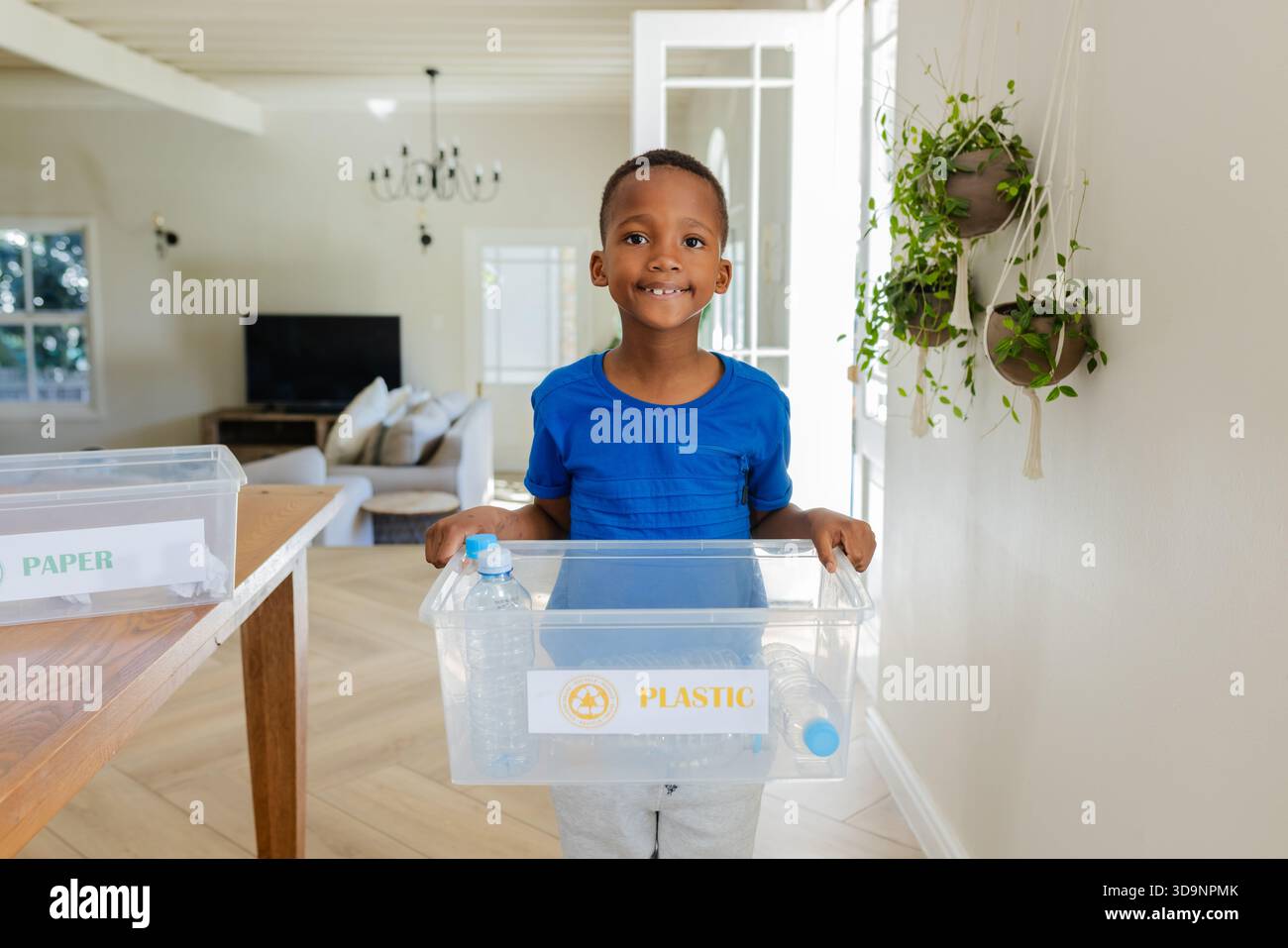 African American child boy standing in modern home living area holding clear bin labeled PLASTIC Stock Photo