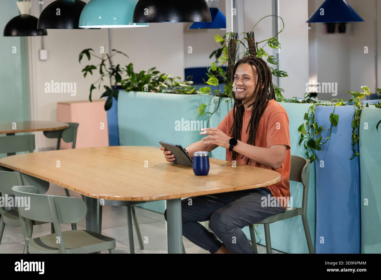 African-American man in orange tee sitting at table in lounge using tablet with thermos, copy space Stock Photo