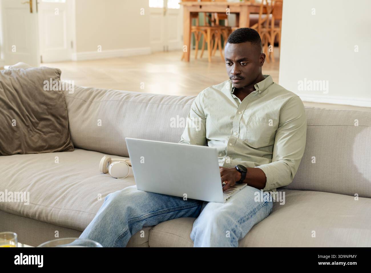African American man working on laptop wearing green shirt, jeans on gray sofa, copy space Stock Photo