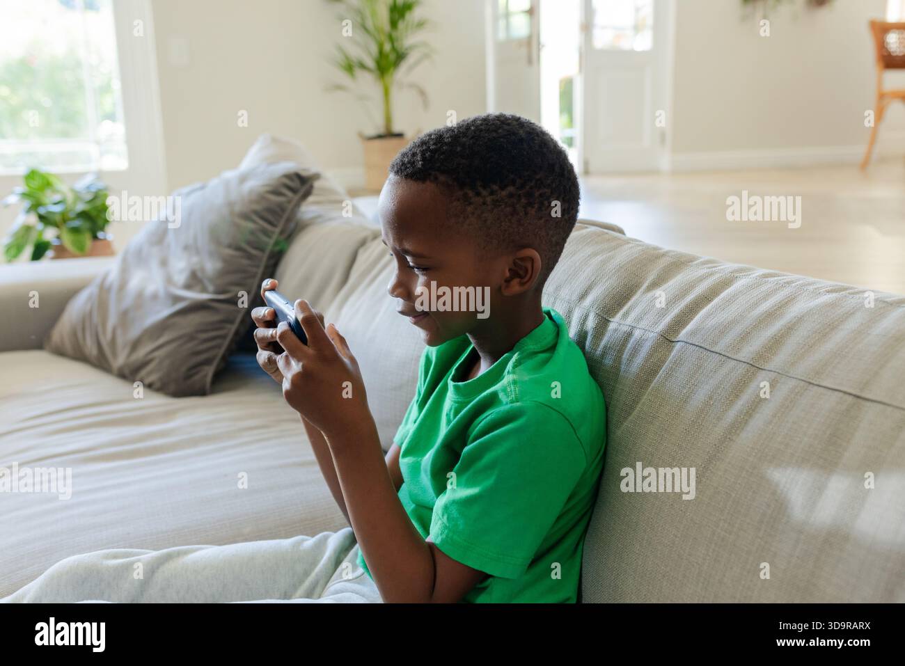 African American child wearing bright green tee sitting on gray sofa holding handheld device Stock Photo