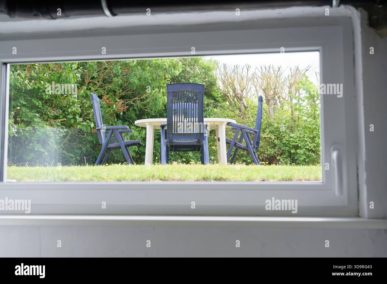 Three black plastic chairs and a small table sit outdoors on grass, surrounded by green bushes, seen through a plastic window Stock Photo