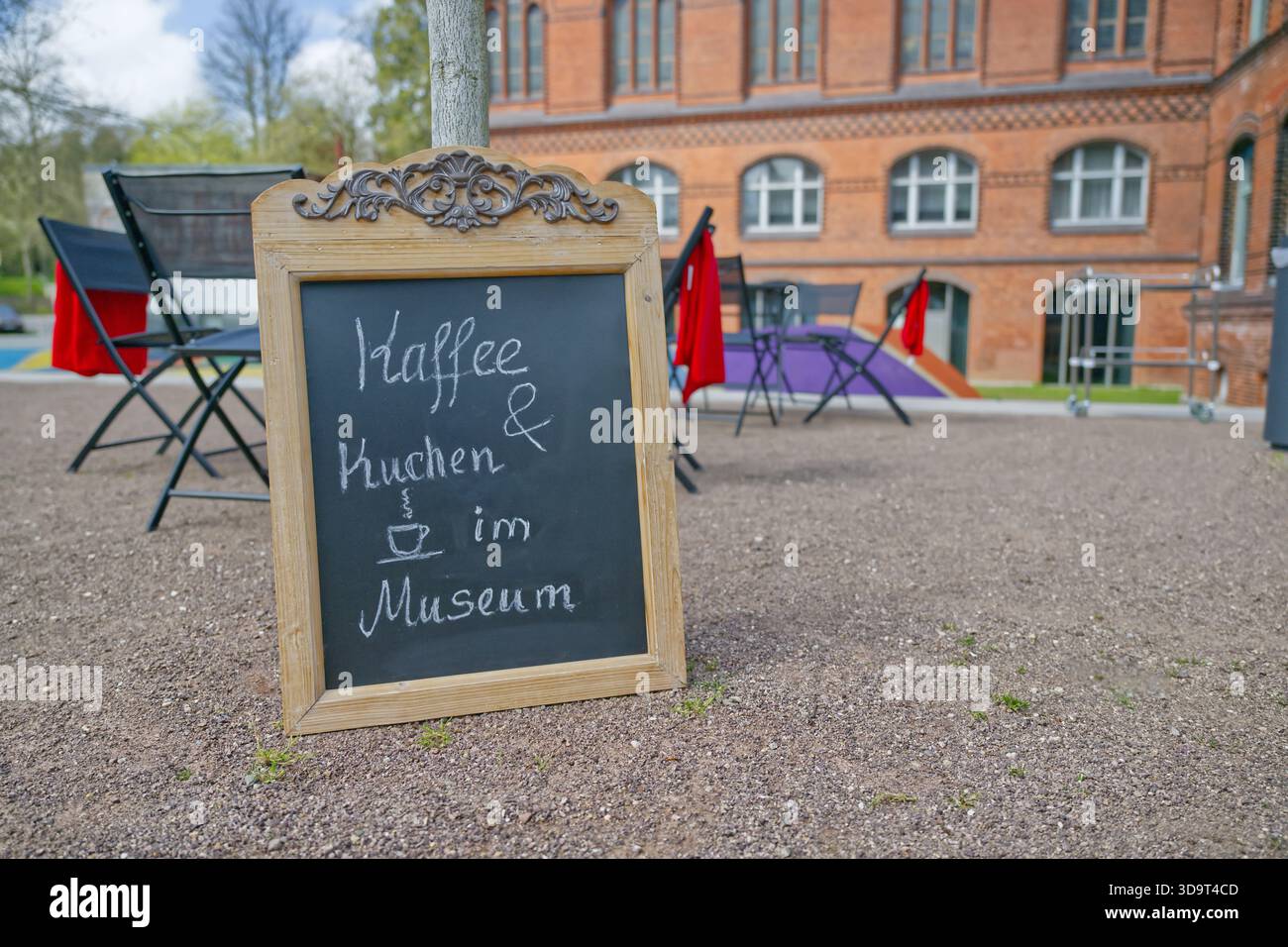 A wooden chalkboard sign displaying Kaffee und Kuchen im Museum at Museumsberg in Flensburg in a scenic outdoor setting with chairs and a museum build Stock Photo