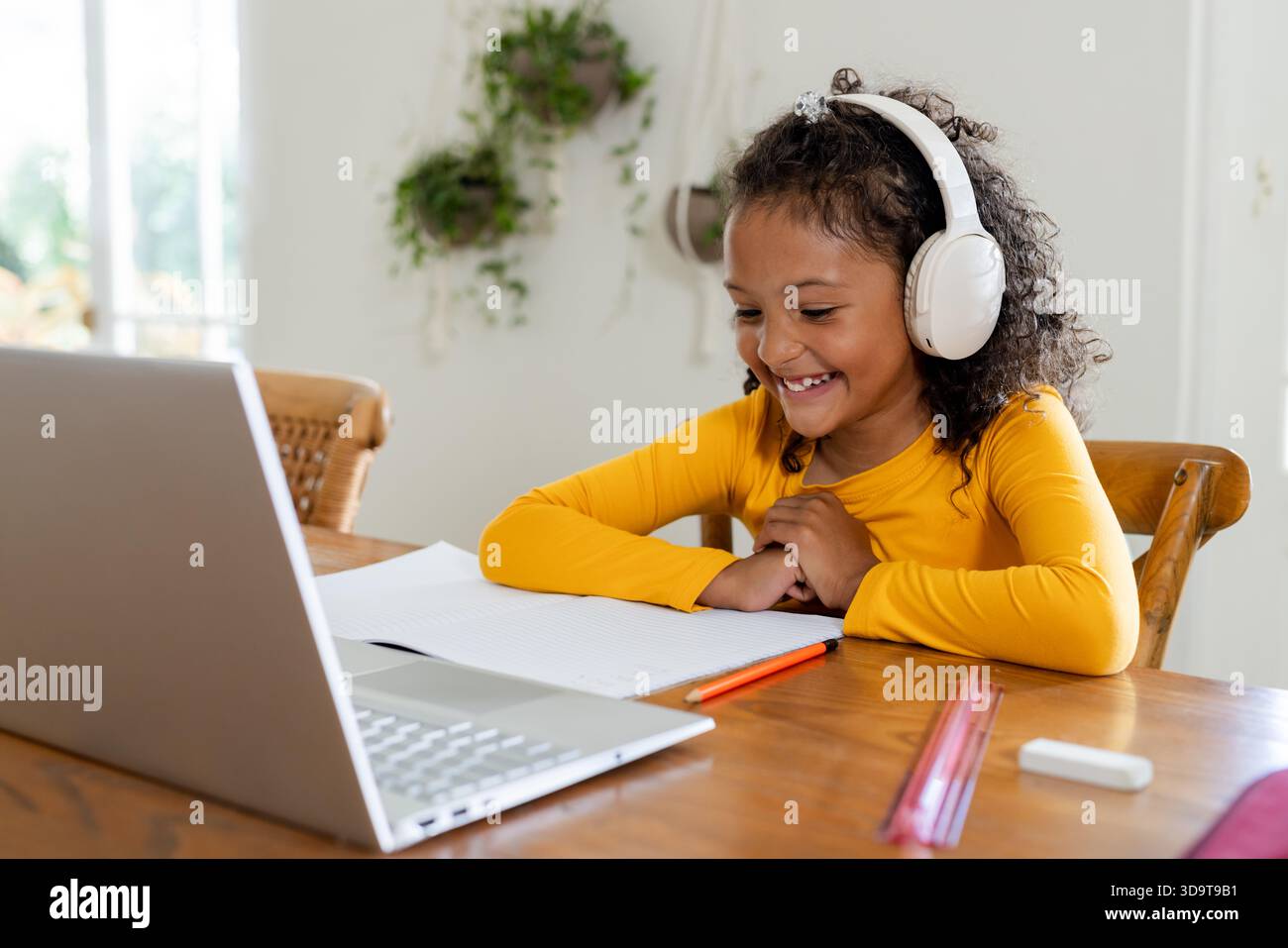 African American child sitting at wooden table at home wearing headphones and studying on laptop Stock Photo