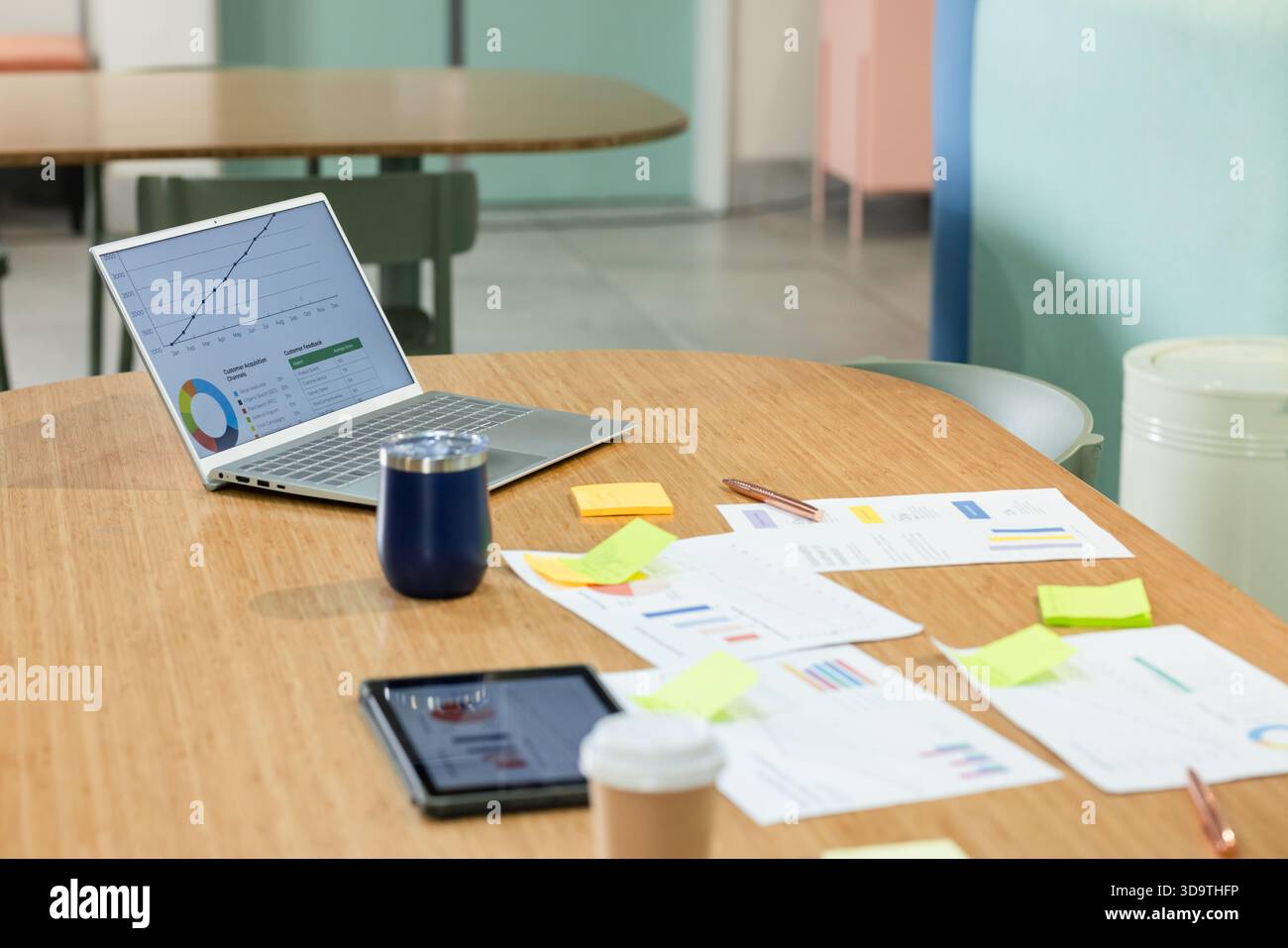 Wooden oval table is holding silver laptop, printed charts, sticky notes, cups in office Stock Photo