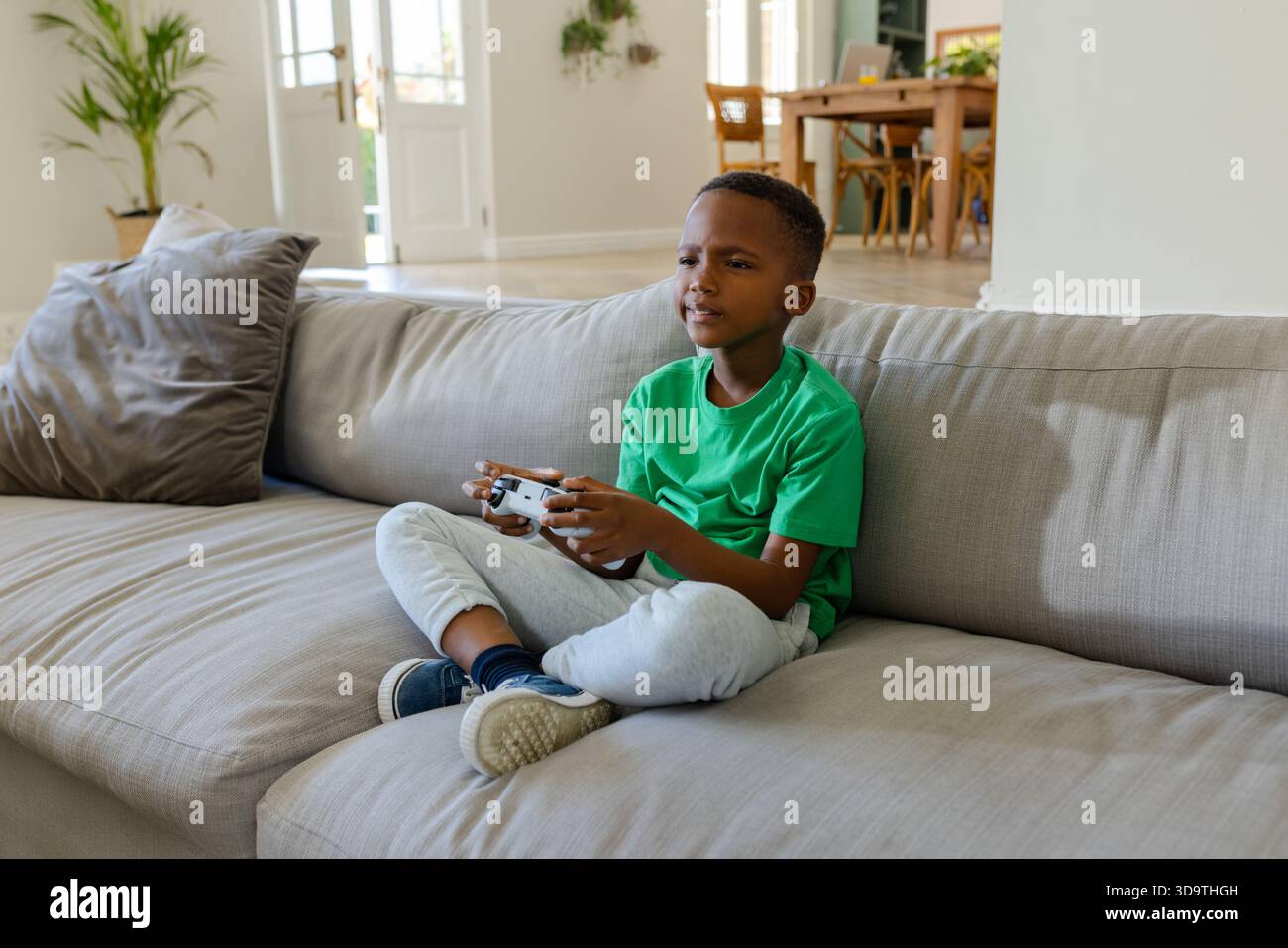 African American green-shirted boy playing on gray sofa at home with white controller, copy space Stock Photo