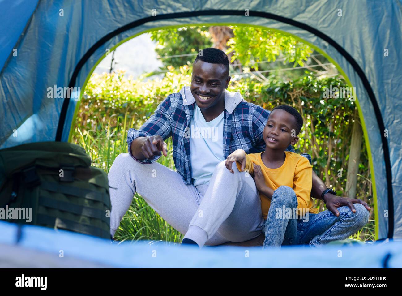 African American man and child sitting at blue tent entrance, man pointing, wearing plaid, backpack Stock Photo