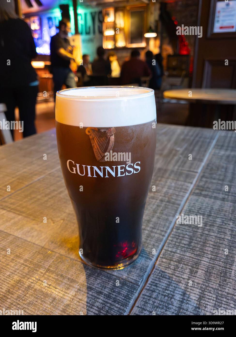 A pint of Guinness in a Guinness branded glass on a table in the lively neighbourhood pub of Pug Uglys, Belfast, NI. Guinness is an Irish Dry Stout - Smartphone Captured Stock Image