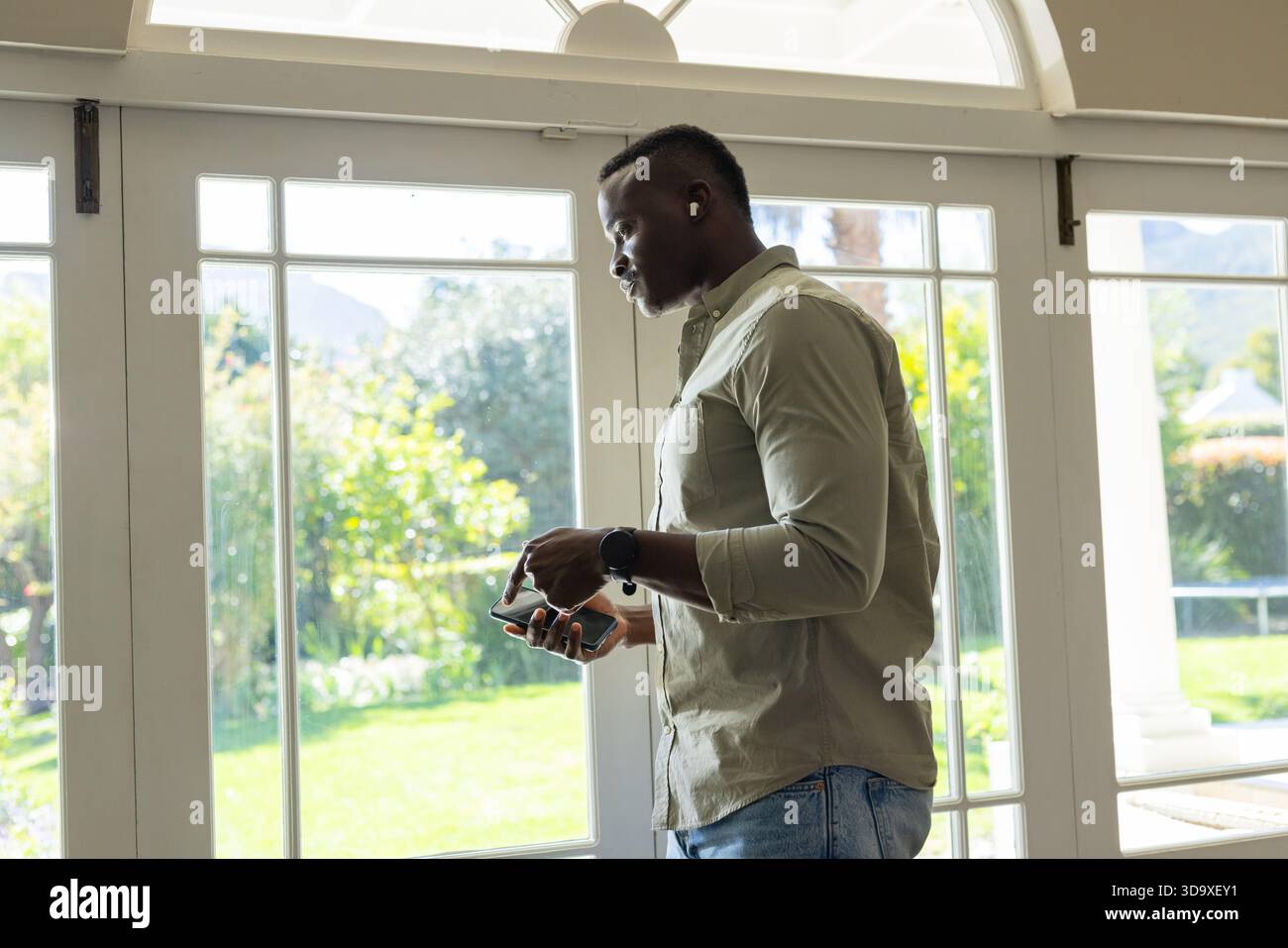 Man is standing by glass French doors and checking phone with earbuds and smartwatch, backyard view Stock Photo
