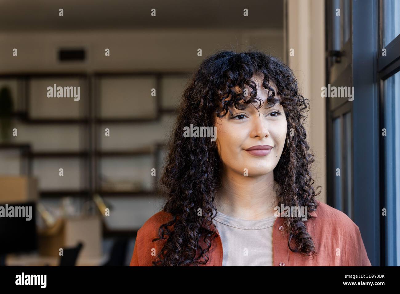 African American woman standing near large window in office space beside shelves boxes workstations Stock Photo