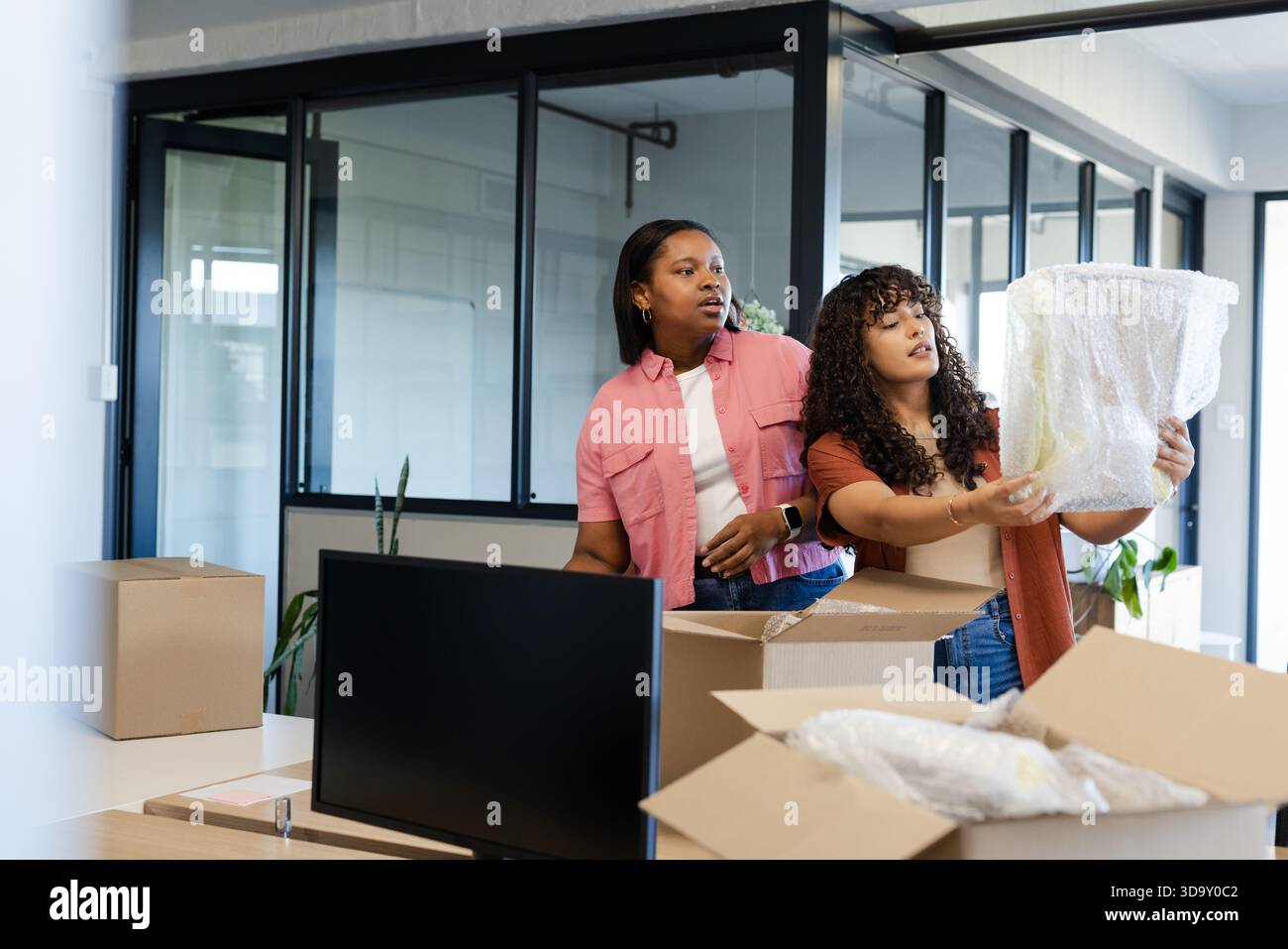 African American female coworkers unpacking bubble-wrapped decor from boxes in office with monitor Stock Photo