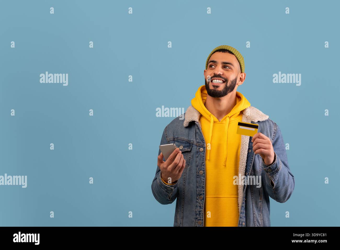 Man Wearing Yellow Hoodie and Denim Jacket Smiles While Holding Credit Card and Smartphone Against Blue Background Stock Photo