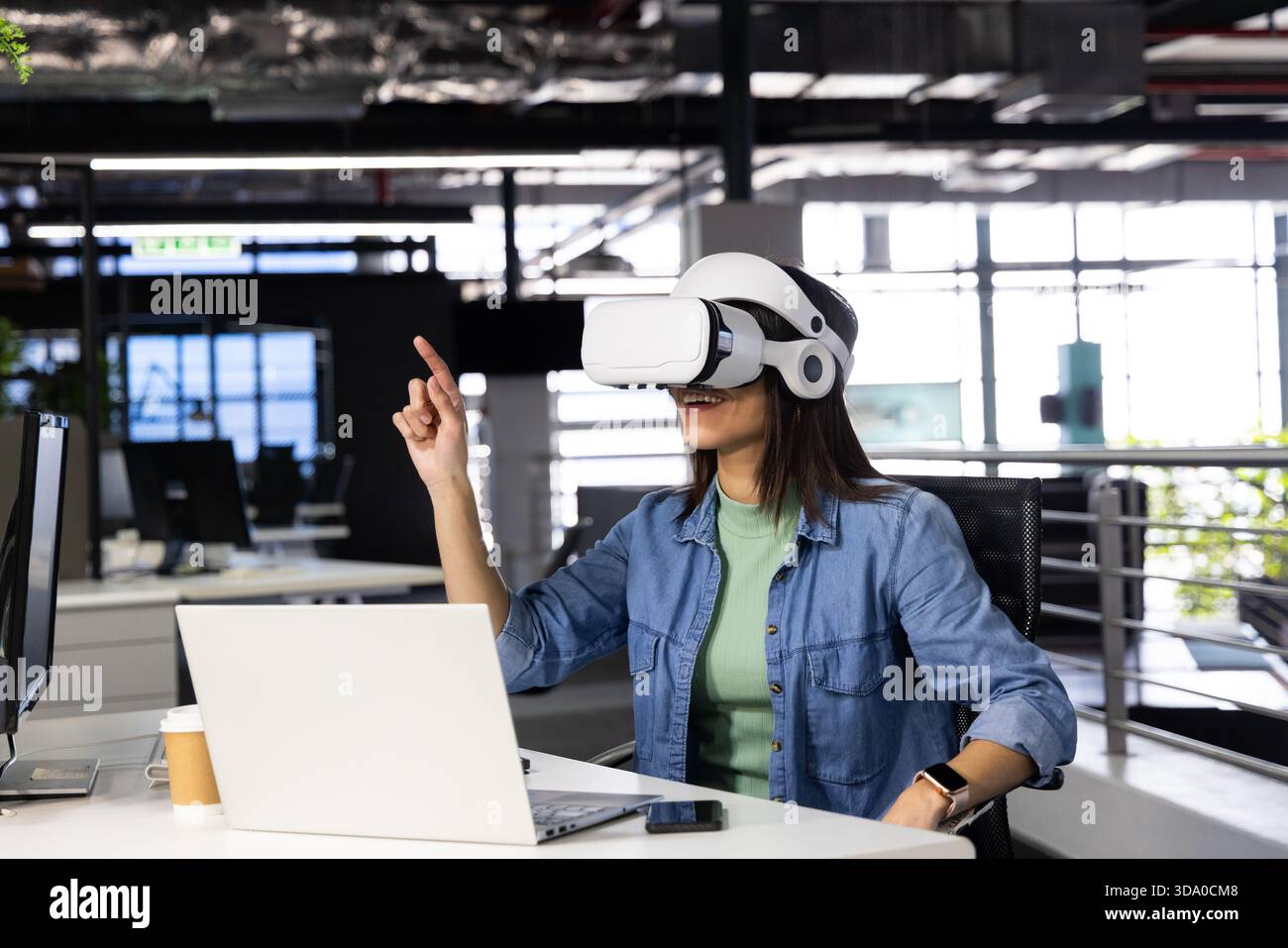 VR headset resting on desk near windows revealing greenery in office, with laptop and coffee cup Stock Photo