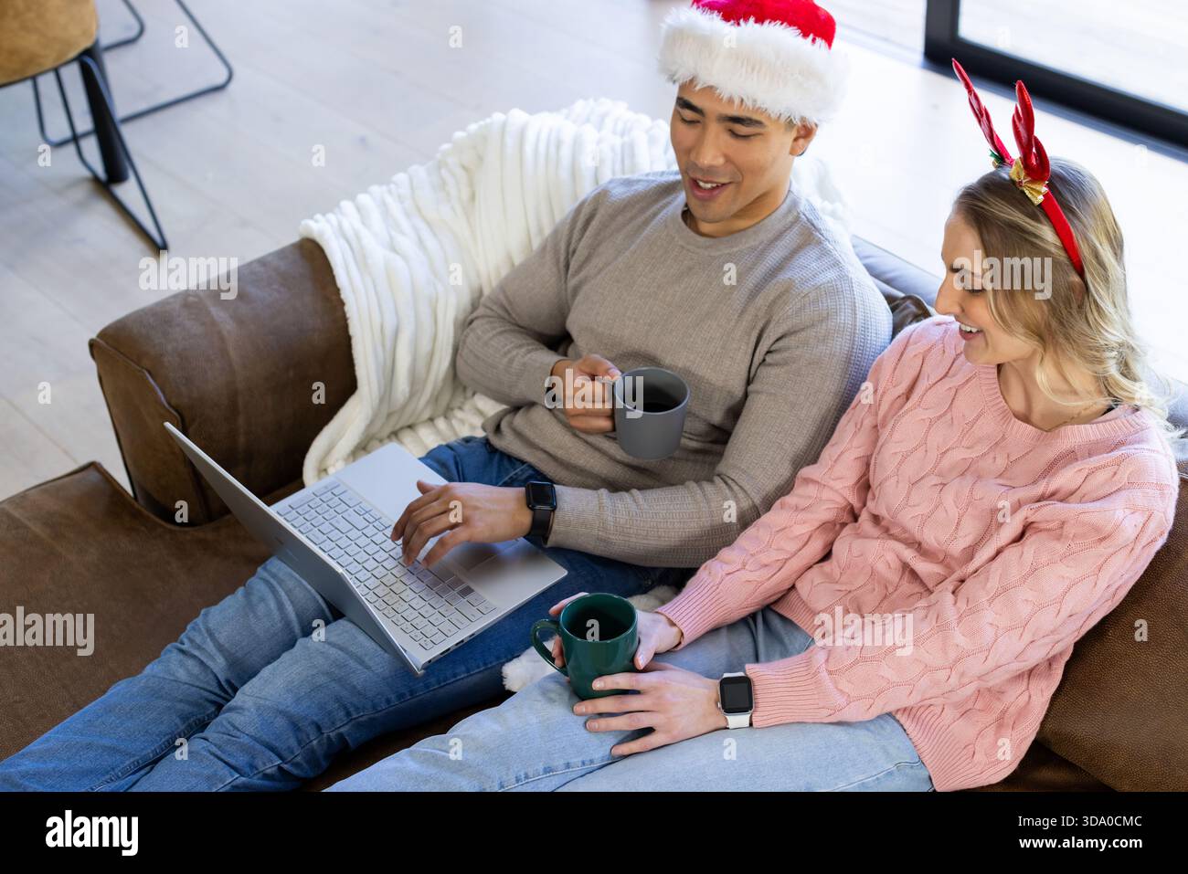 Diverse couple wearing Santa hat and antlers, sitting on couch at home holding mugs, using laptop Stock Photo