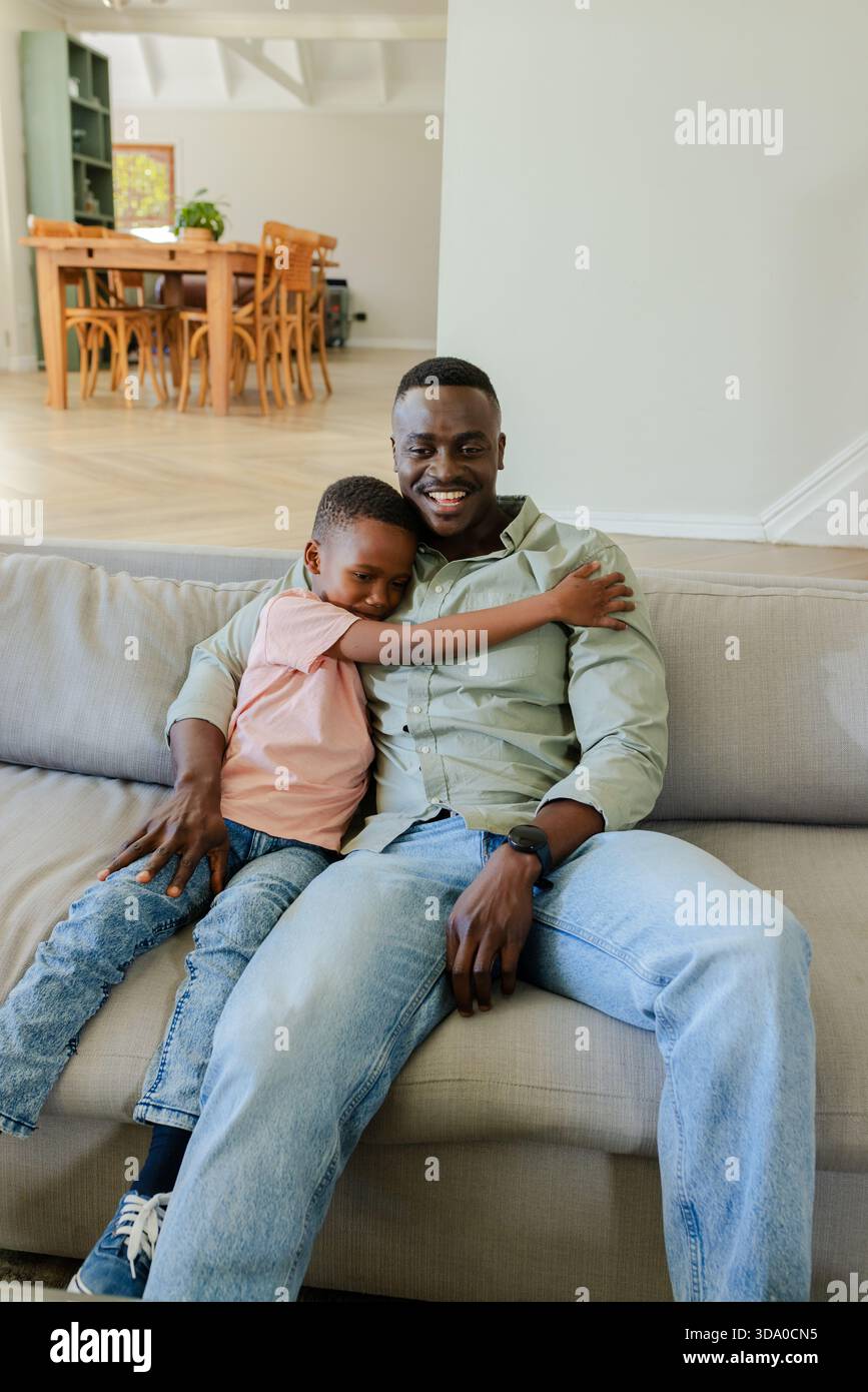 African American father and son sitting on light-colored sofa in living room with potted plant Stock Photo