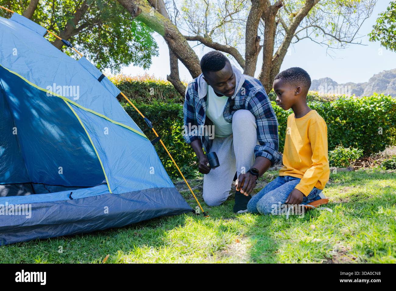 African American father and child kneeling securing blue dome tent with mallet and stakes on lawn Stock Photo