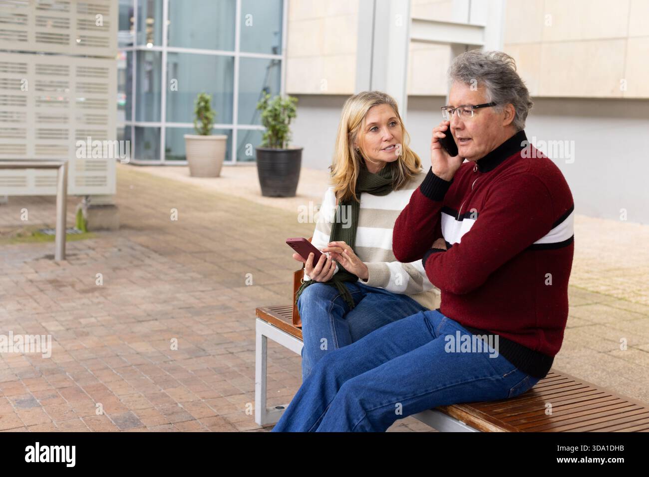Senior couple sitting on bench in courtyard, woman holding smartphone and man talking on phone Stock Photo