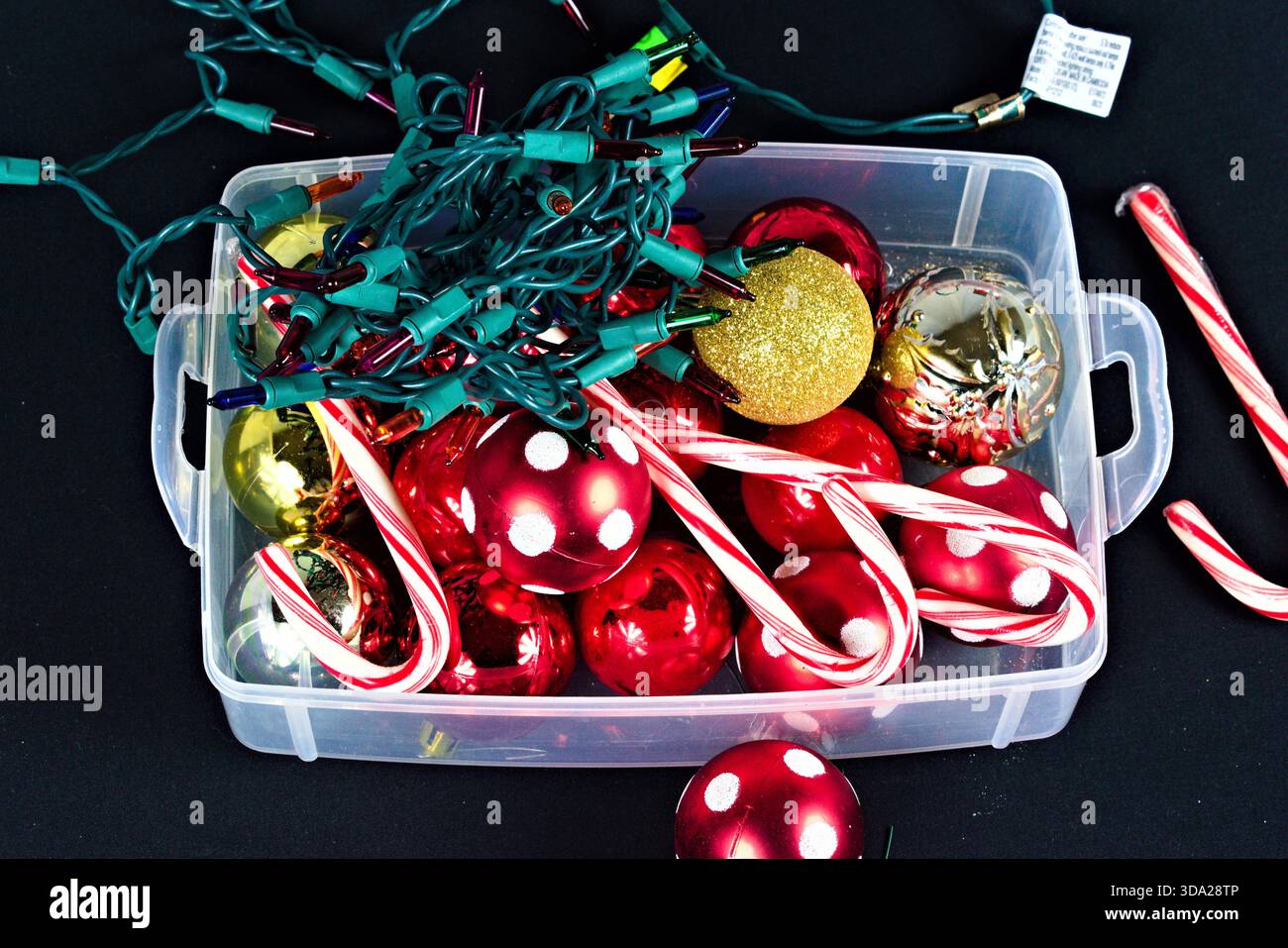 A clear plastic box with different colored Christmas ornaments, a string of lights, and some candy canes against a black background. Stock Photo
