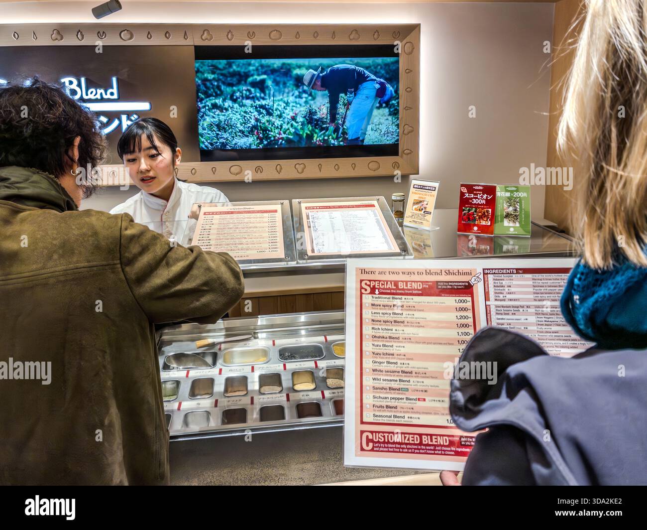 A staff member helps a customer mix a personal shichimi blend from small trays of individual spices at the Yawataya Isogoro (八幡屋礒五郎) flagship store on Daimon-cho in Nagano City. The counter menu lists blend options such as extra spicy, mild, yuzu citrus or white sesame, starting at 1,000 yen. Stock Photo
